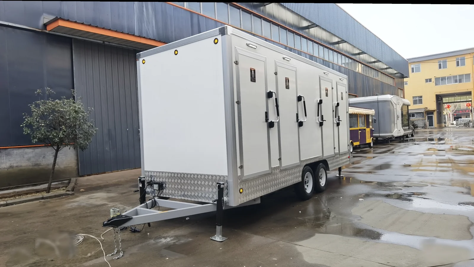 Front angle view of a 4 stall restroom trailer with towing frame, dual axle chassis, stabilizer jacks, and factory yard background.
