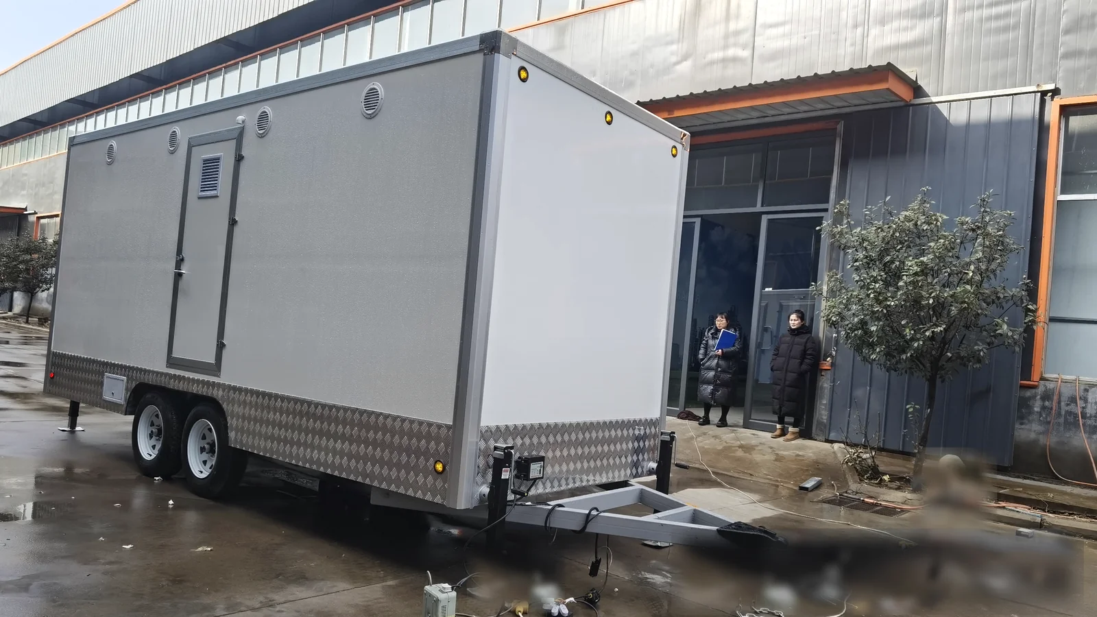 Front service side view of a 4 stall restroom trailer with towing frame, side panel, dual axle chassis, and factory yard background.