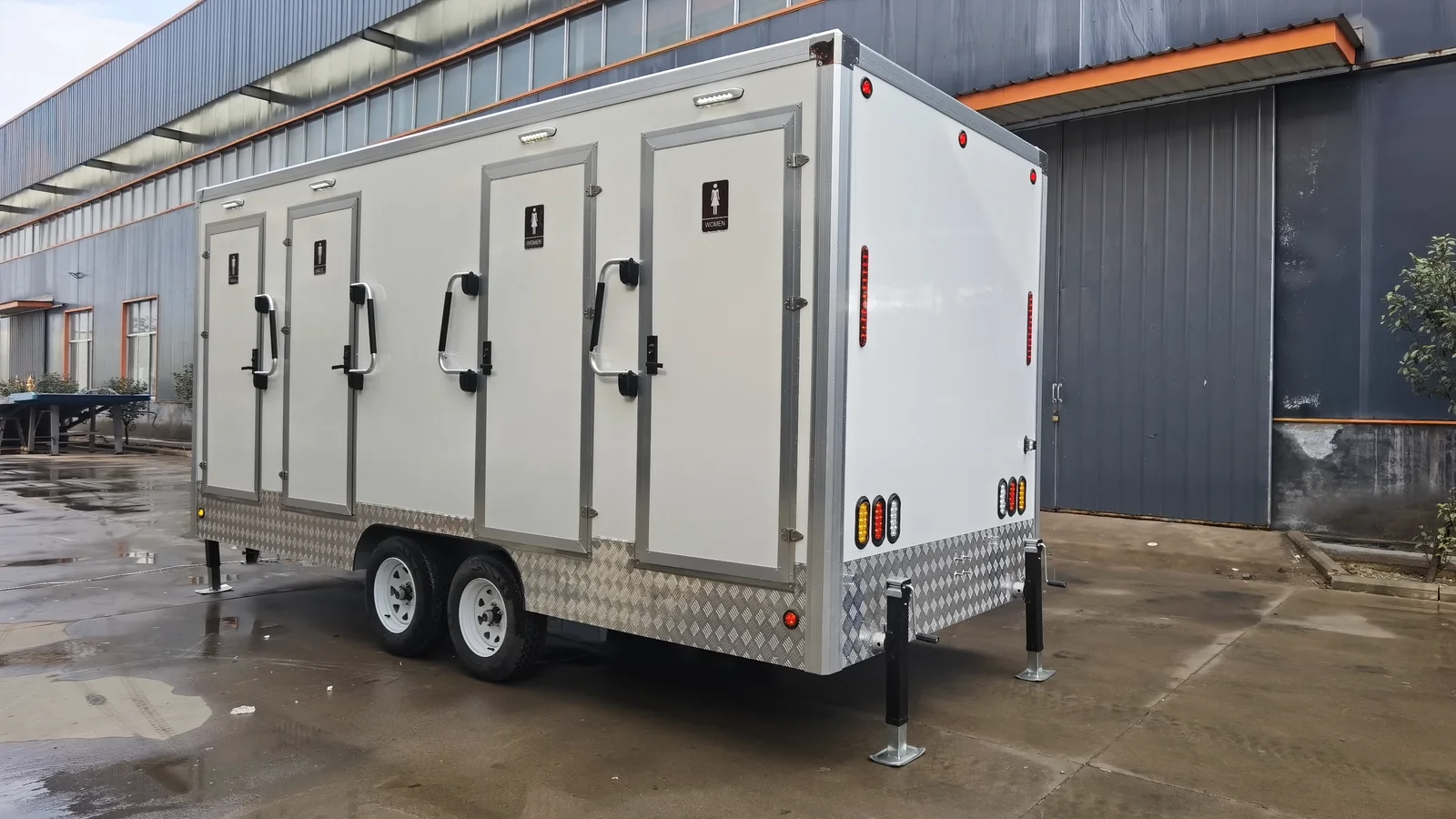Rear angle view of a 4 stall restroom trailer with rear lights, side doors, stabilizer legs, and factory production background.