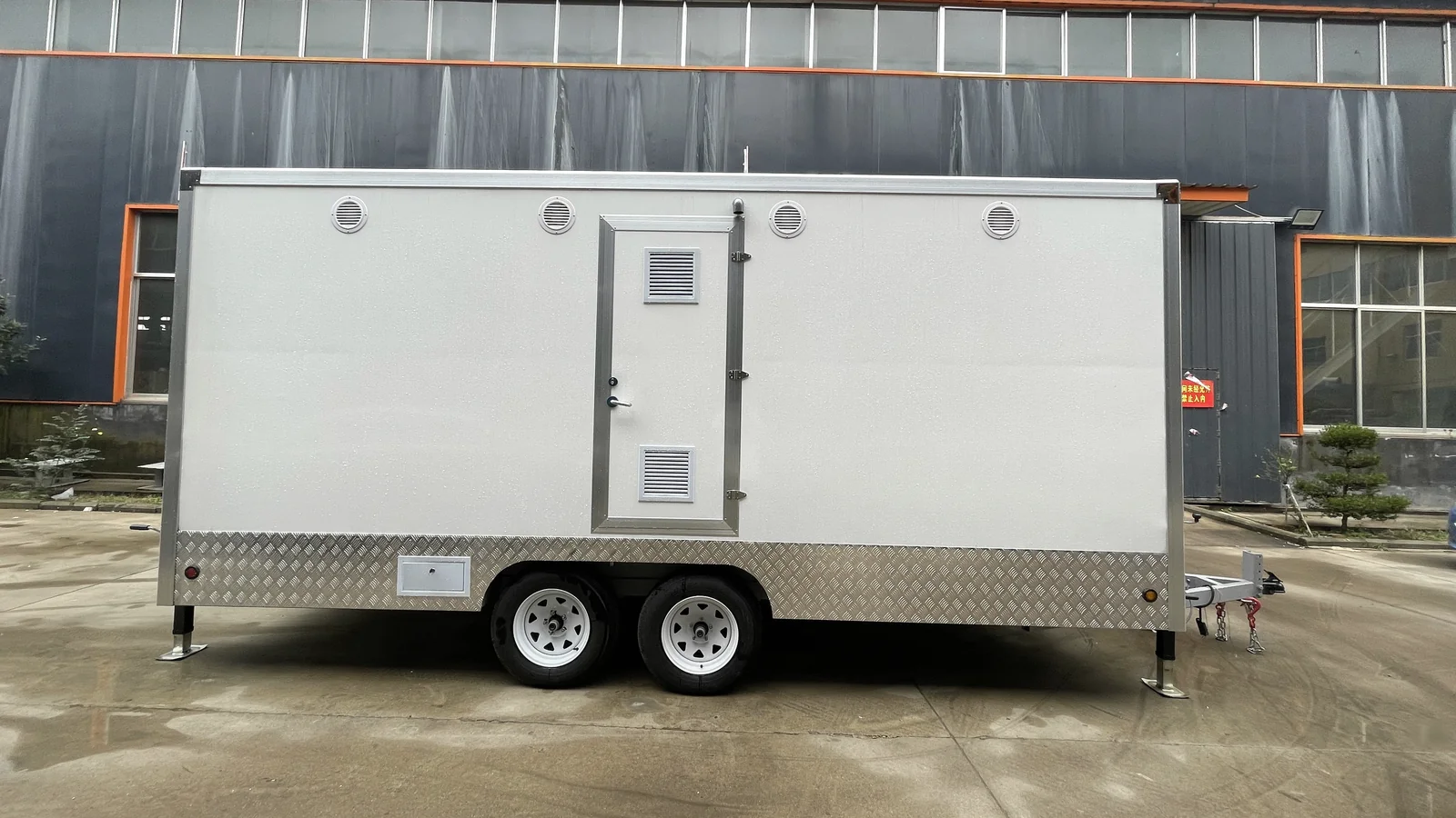 Service side view of a 4 stall restroom trailer showing side panels, ventilation, chassis, stabilizer legs, and towing-ready structure.